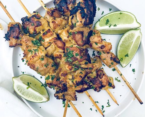 Chicken satay skewers and lime wedges on a white platter.