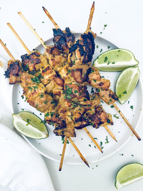 Chicken satay skewers and lime wedges on a white platter.