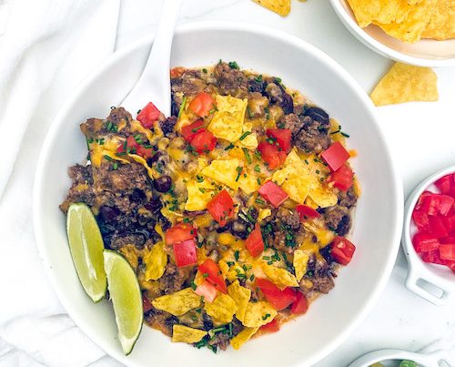 Taco casserole in a white bowl with corn chips and lime wedges.