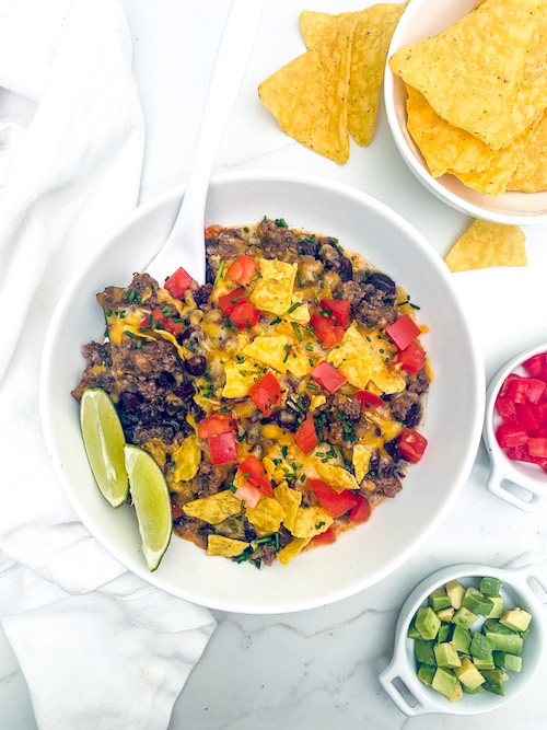 Taco casserole in a white bowl with corn chips and lime wedges.