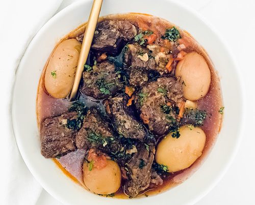 A white bowl and gold spoon with pot roast and potatoes.