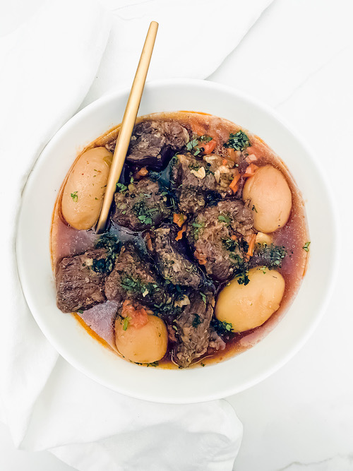 A white bowl and gold spoon with pot roast and potatoes.