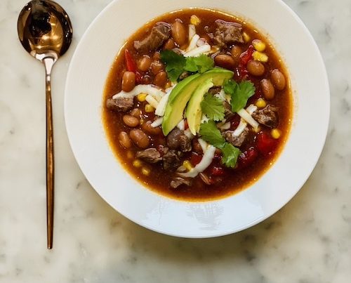 A white bowl filled with spicy beef and bean stew topped with avocado.