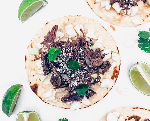 Barbacoa beef with onions on tortillas on a white surface.