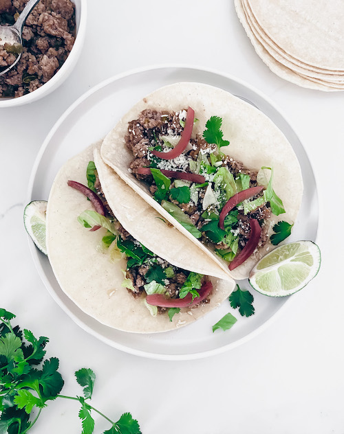 Two ground pork tacos on a white plate with cilantro and lime.