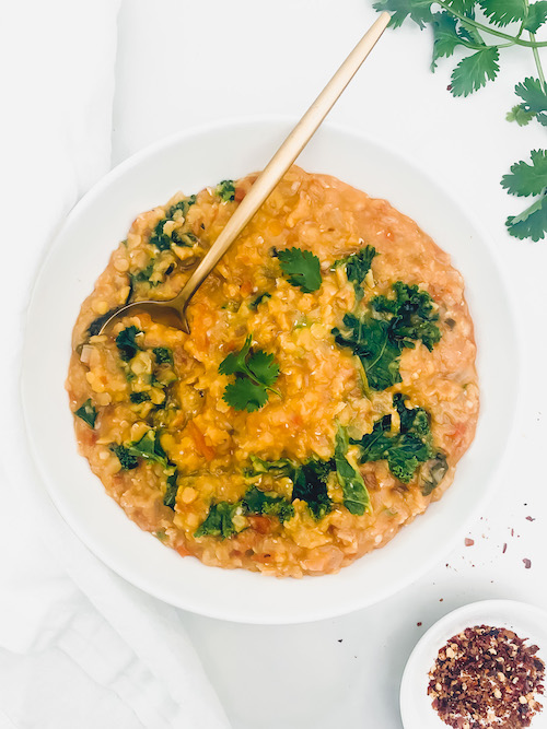 Red Lentil + Kale Daal in a white bowl with gold serving spoon.