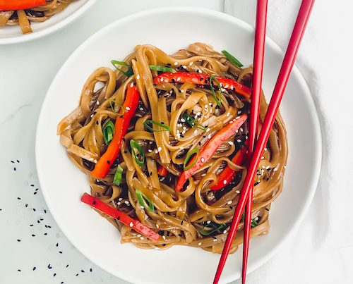 A white bowl filled with drunken noodles and red peppers with a pair of chopsticks.