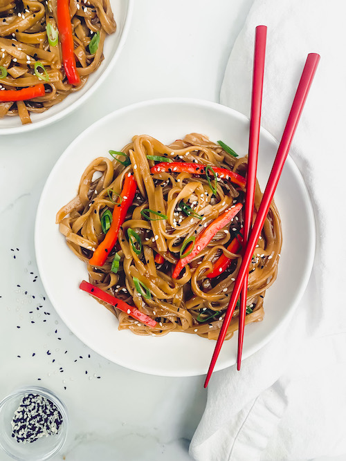 A white bowl filled with drunken noodles and red peppers with a pair of chopsticks.