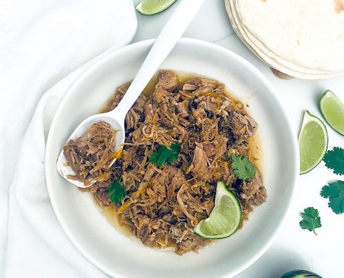 Shredded pork fajitas with lime in a white serving bowl with spoon.
