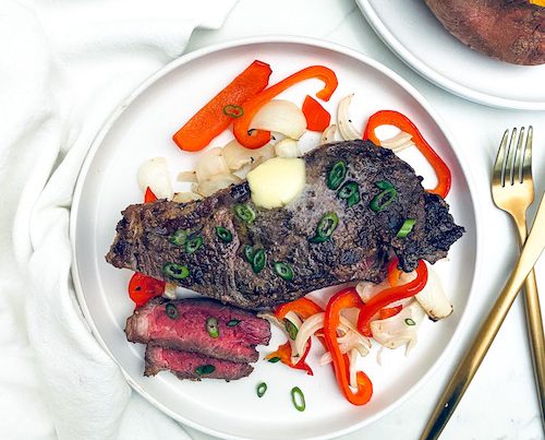 Strip steak that has been air fried resting on a plate with onions and peppers.