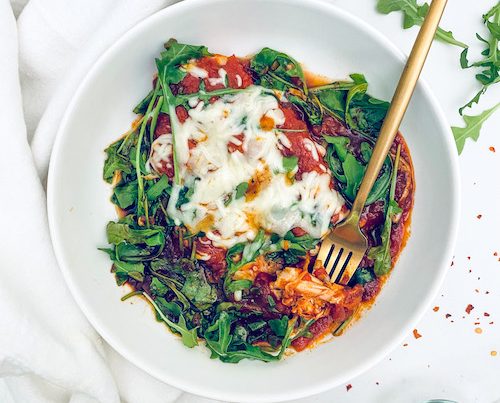 Mozzarella topped chicken with arugula in a white bowl.