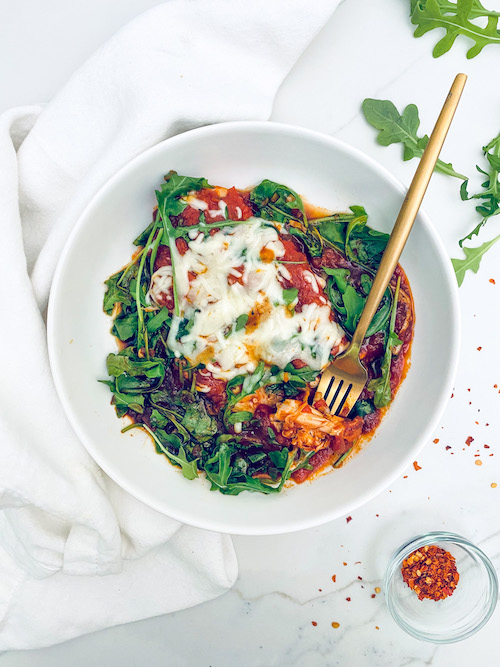 Mozzarella topped chicken with arugula in a white bowl.