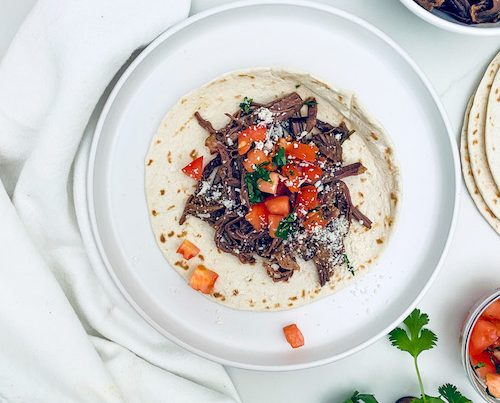 A small flour tortilla on a white plate topped with shredded steak.