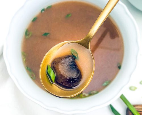 A mushroom on a gold spoon over a bowl of clear onion soup.