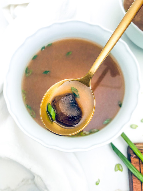 A mushroom on a gold spoon over a bowl of clear onion soup.