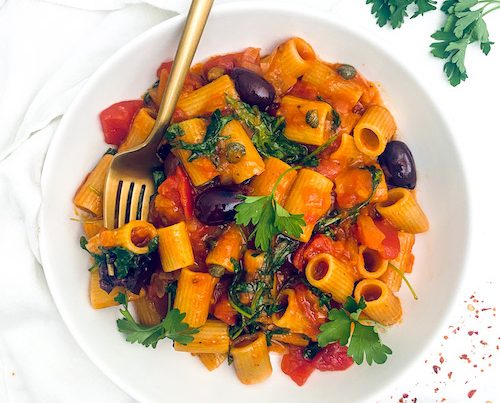 Rigatoni in sauce with olives and tomatoes in a white bowl.