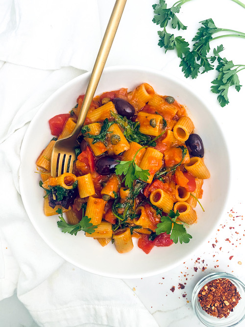 Rigatoni in sauce with olives and tomatoes in a white bowl.