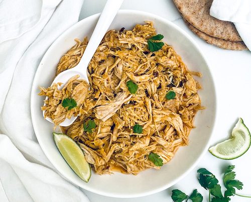 Shredded tandoori chicken in a white bowl with lime wedges.